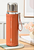 A vintage-style orange water bottle with a stainless steel cap and wrist strap, shown on a tabletop with decorative flowers and books in the background. The image includes callout graphics on the left describing features like the 630ml capacity, leakproof seal, and flip-top lid.
