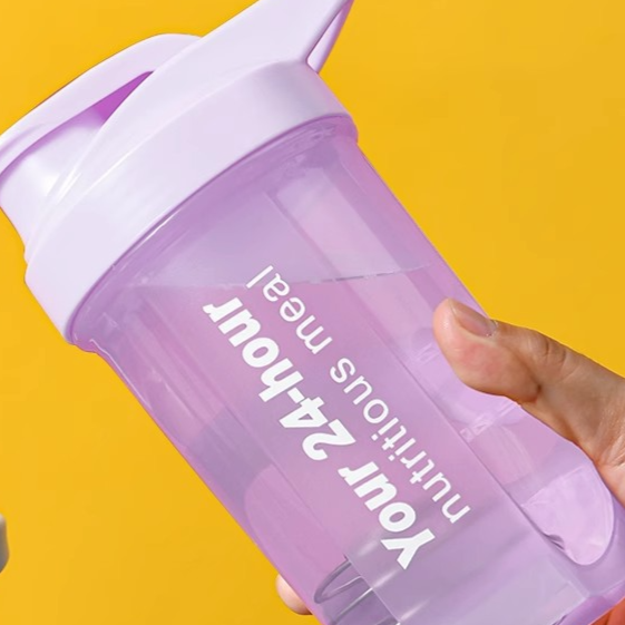 A translucent purple shaker bottle with a lavender lid, held by a hand against a solid yellow background. The bottle features white text that reads 'Your 24-hour nutritious meal'.