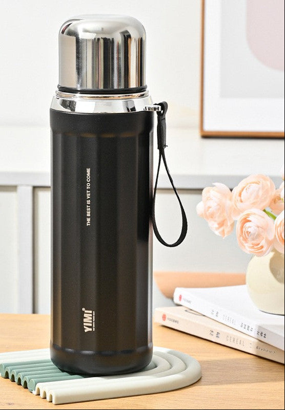 A black 630ml stainless steel vacuum flask with a silver cap and a black wrist strap, displayed on a light-colored coaster on a wooden table. The background is a bright, modern room with pink roses in a vase.