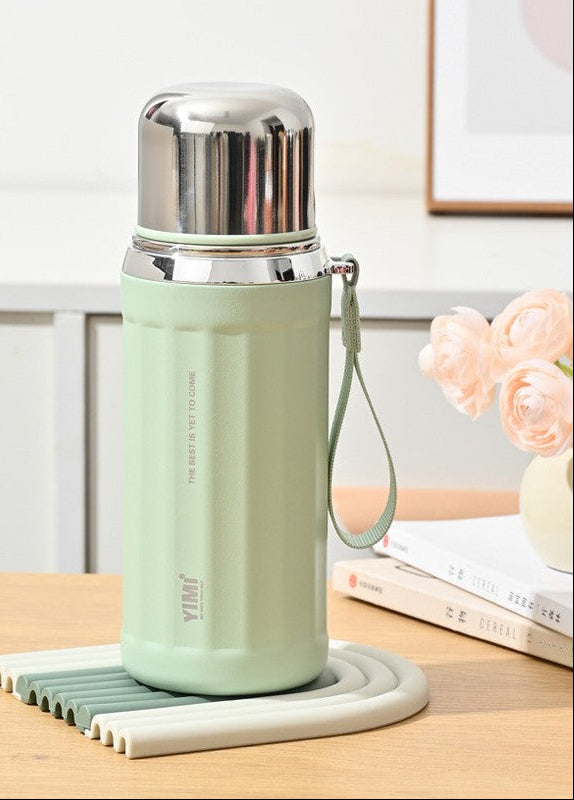 A 530ml spruce green insulated water bottle with a stainless steel lid and carrying loop, displayed on a matching silicone coaster on a wooden table. The image includes feature callout graphics on the left and a soft home background with books and flowers on the right.