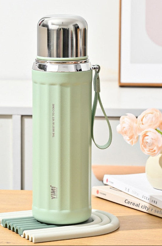 A mint green insulated water bottle with a stainless steel lid and wrist strap, shown on a tabletop with decorative flowers and books in the background. The image includes feature callout graphics on the left and text reading 'Spruce green' and '630ml'.