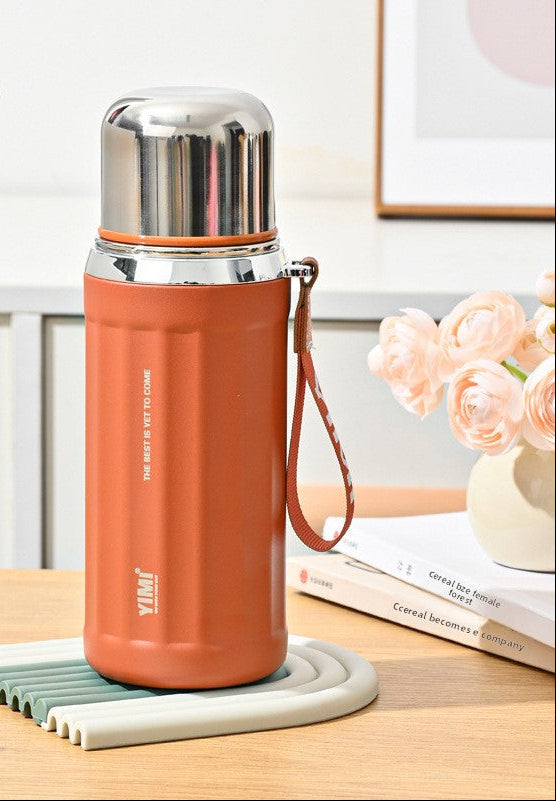 A 530ml vintage orange stainless steel water bottle with a carrying strap, displayed on a coaster on a wooden table with decorative books and flowers in the background. The image includes feature callout graphics on the left.