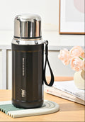A black 530ml stainless steel water bottle with a silver cap and wrist strap, shown on a tabletop with decorative flowers and books in the background. The image includes callout graphics on the left describing features like the straw, warm hand warmers, and leakproof seal.