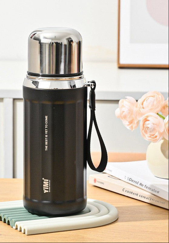 A black 530ml stainless steel water bottle with a silver cap and wrist strap, shown on a tabletop with decorative flowers and books in the background. The image includes callout graphics on the left describing features like the straw, warm hand warmers, and leakproof seal.